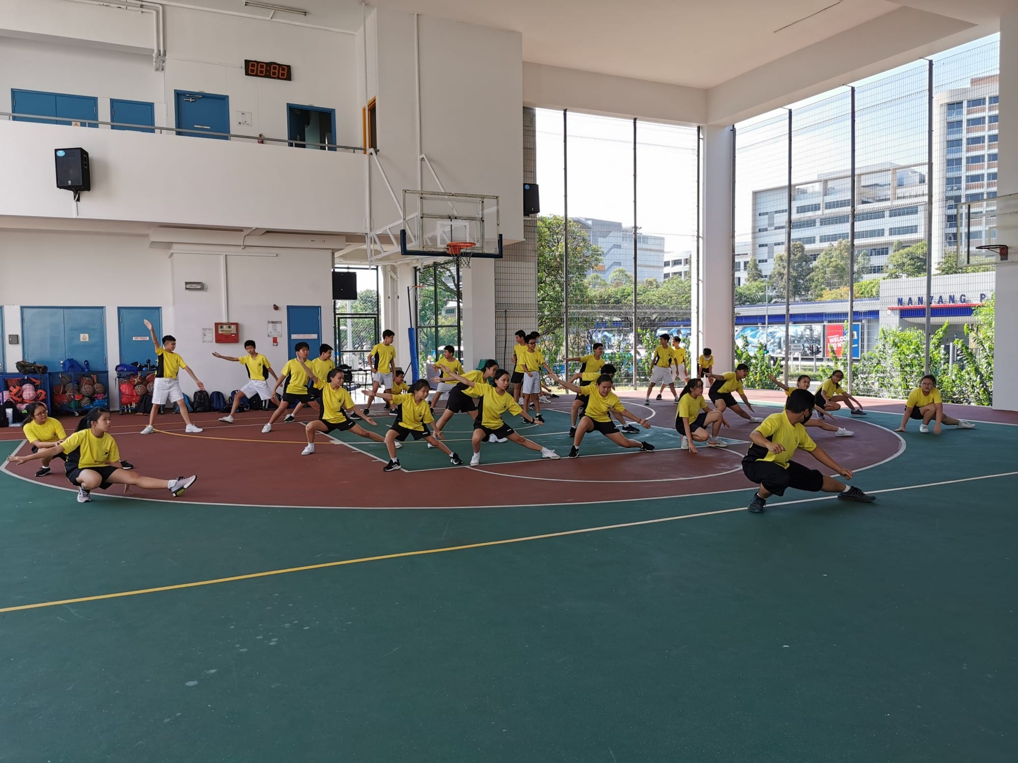A large group of students in yellow shirts stretching and exercising together on an indoor basketball court.